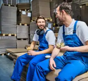Trabajadores en fábrica comiendo, pausa laboral en almacén, empleados felices en entorno industrial, descanso en línea de producción, ambiente laboral positivo y colaborativo, trabajadores con comida en mano, camaradería en el trabajo. – SoyMigrante.com REVISTA