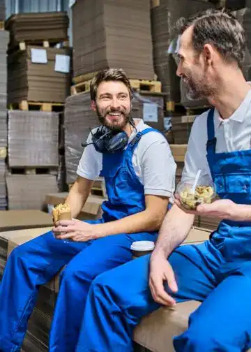 Trabajadores en fábrica comiendo, pausa laboral en almacén, empleados felices en entorno industrial, descanso en línea de producción, ambiente laboral positivo y colaborativo, trabajadores con comida en mano, camaradería en el trabajo. – SoyMigrante.com REVISTA