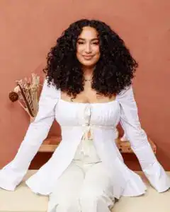 Retrato de mujer con cabello rizado oscuro, sonriendo y vestida con atuendo blanco, en un entorno acogedor, promoviendo temas de migrantes y cultura en SoyMigrante.com REVISTA. – SoyMigrante.com REVISTA