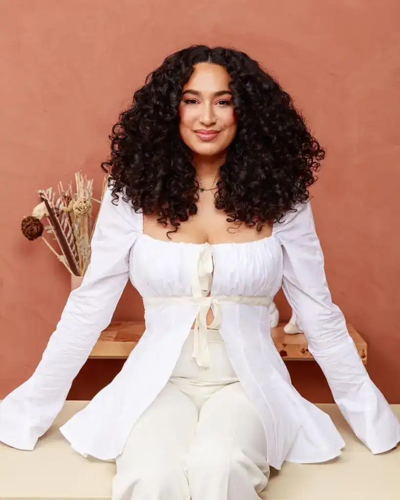 Retrato de mujer con cabello rizado oscuro, sonriendo y vestida con atuendo blanco, en un entorno acogedor, promoviendo temas de migrantes y cultura en SoyMigrante.com REVISTA. – SoyMigrante.com REVISTA