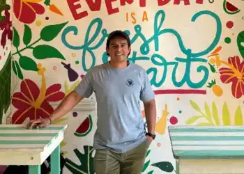 Personaje sonriente en ambiente colorido y tropical con mural que dice "Everyday is a fresh start", promoviendo bienestar y positivismo para migrantes.