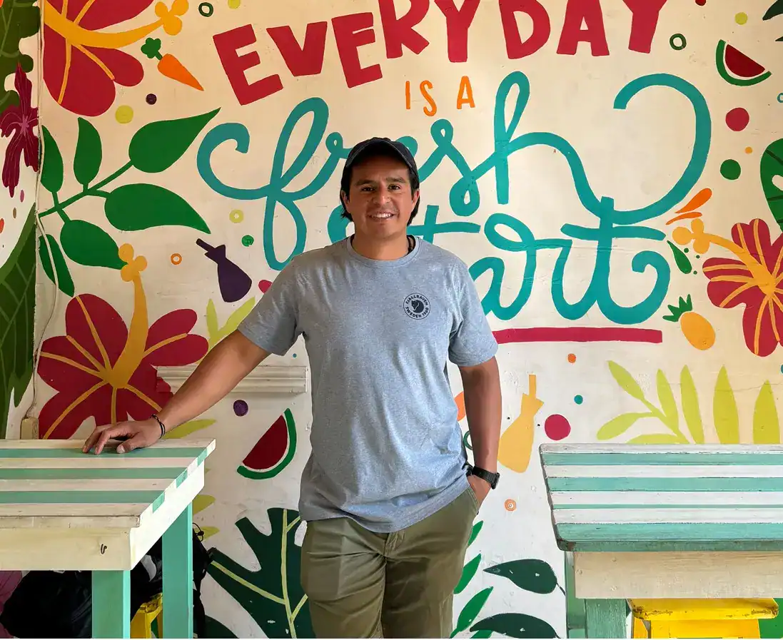 Personaje sonriente en ambiente colorido y tropical con mural que dice "Everyday is a fresh start", promoviendo bienestar y positivismo para migrantes. – SoyMigrante.com REVISTA