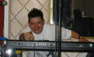 DJ sonriendo y señalando, en un evento musical, en un ambiente con decoración moderna, transmitiendo alegría y cultura, promocionando diversidad y comunidad en la revista SoyMigrante.com REVISTA. – SoyMigrante.com REVISTA