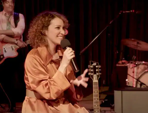 SoyMigrante.com REVISTA - Artista mujer cantando con guitarra en concierto en un escenario con fondo de cortinas rojas. – SoyMigrante.com REVISTA