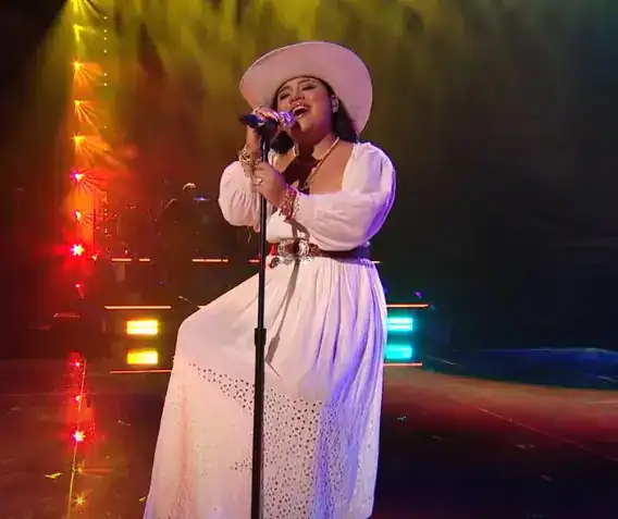Voz femenina cantando en vivo, vestida con traje elegante y sombrero grande en escenario de concierto, reflejando talento y pasión musical.