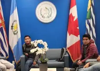 Relevancia de la imagen en relación con la revista SoyMigrante.com REVISTA y su temática de migración, integración y derechos de migrantes en Guatemala. La foto muestra un grupo diverso de jóvenes en una reunión oficial en un entorno institucional con banderas y logotipos oficiales.