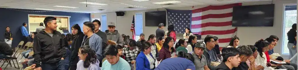 Diversidad migrante en evento comunitario con bandera estadounidense en el fondo.