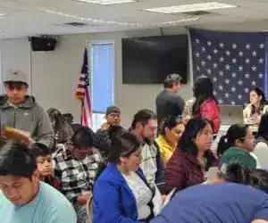Diversidad migrante en evento comunitario con bandera estadounidense en el fondo.