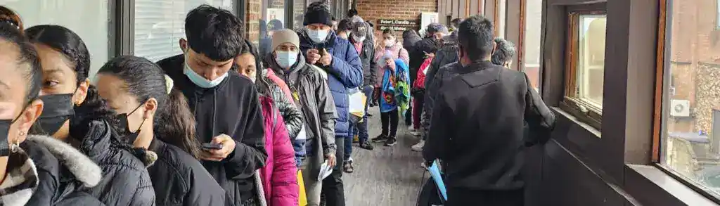 Larga fila de personas con mascarillas esperando en una oficina o agencia, ilustrando temas de migrantes, movilidad y atención al cliente.