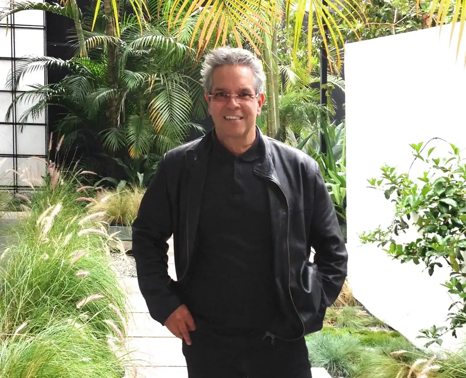 Hombre sonriendo en un entorno verde y tropical, con plantas y palmeras, ilustrando el contenido de la revista SoyMigrante.com REVISTA, que promueve historias y noticias sobre migración, integración y derechos en América Latina y el mundo. – SoyMigrante.com REVISTA