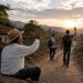 Older man in a straw hat sits on the ground and waves to two hikers on a dirt road at sunset in a rural landscape.