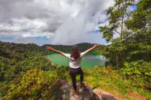 Espejo de agua en medio de la selva en un paisaje natural, mujer con los brazos abiertos disfrutando vista panorámica en un destino ecoturístico, turismo de naturaleza y aventuras en Latinoamérica. – SoyMigrante.com REVISTA