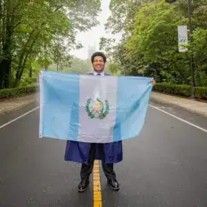Guatemalteco con toga y birrete sosteniendo la bandera de Guatemala en una calle arbolada, celebrando logros académicos y el orgullo de ser migrante guatemalteco en el extranjero. – SoyMigrante.com REVISTA