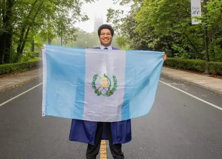 Guatemalteco con toga y birrete sosteniendo la bandera de Guatemala en una calle arbolada, celebrando logros académicos y el orgullo de ser migrante guatemalteco en el extranjero.