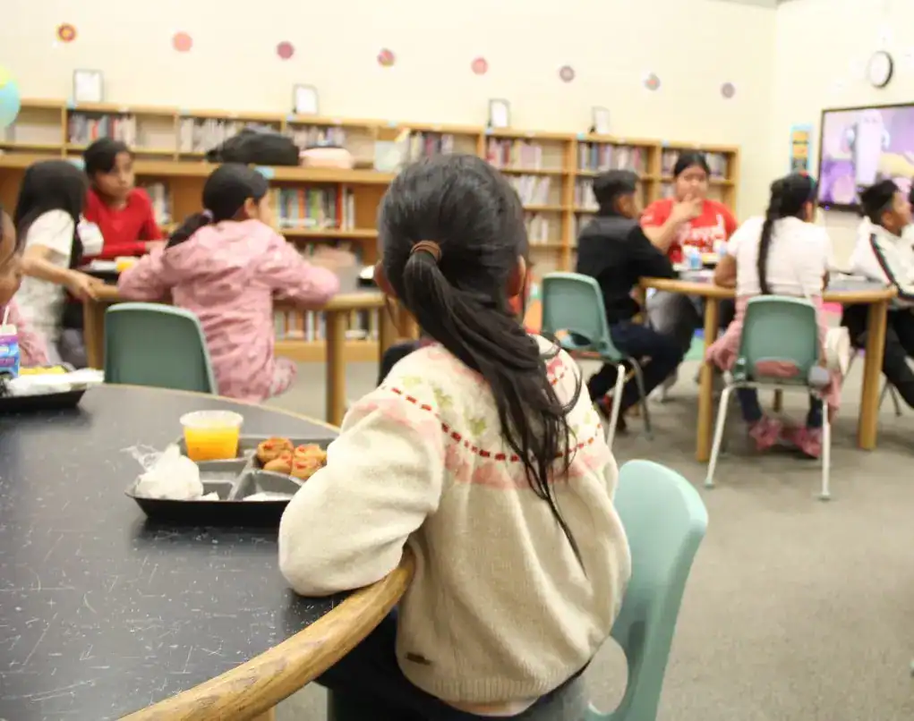 Estudiante migrante en aula de biblioteca, participando en actividades educativas con compañeras para promover la integración y el aprendizaje en un entorno escolar inclusivo. – SoyMigrante.com REVISTA