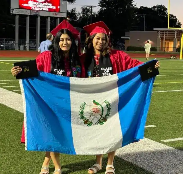 Orgullo y celebración de graduación con la bandera de Guatemala en un evento educativo. Dos jóvenes en toga y birrete muestran su logro académico en un ambiente escolar al aire libre. – SoyMigrante.com REVISTA