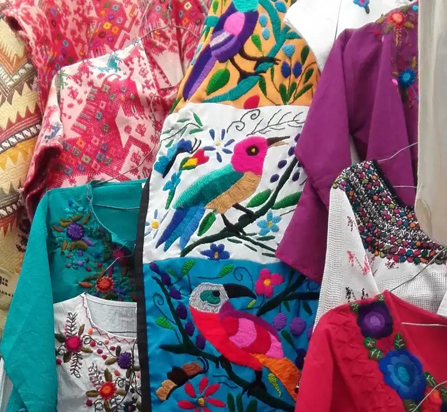 Coloridas prendas con bordados tradicionales y diseños de flora y fauna, ideales para promover la cultura mexicana, el arte textil y la moda étnica, en una exhibición de textiles artesanales. – SoyMigrante.com REVISTA