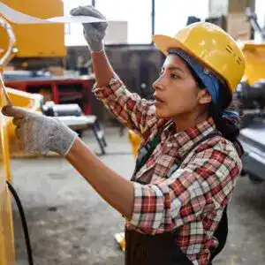 Trabajadora migrante en taller de construcción con casco de seguridad, dirigendo tareas en fábrica, promoviendo la integración laboral y la inclusión de migrantes en el sector industrial en España. – SoyMigrante.com REVISTA