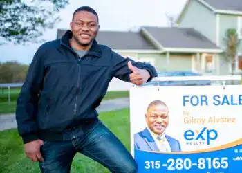 Gilroy Alvarez dfdf – SoyMigrante.com REVISTA Vendedor de bienes raíces mostrando un cartel de propiedad en venta con enfoque en la comunidad migrante en EE. UU. y servicios de inmobiliaria. – SoyMigrante.com REVISTA