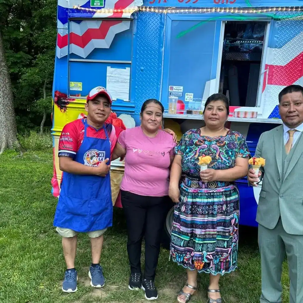 Heladeros y clientes en una feria de comida mexicana, disfrutando de helados artesanales frente a un food truck colorido en un entorno verde y natural.