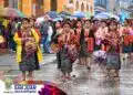 Tradicional desfile folklórico de niñas en San Juan Sacatepequez con vestimenta típica y paraguas en día lluvioso.