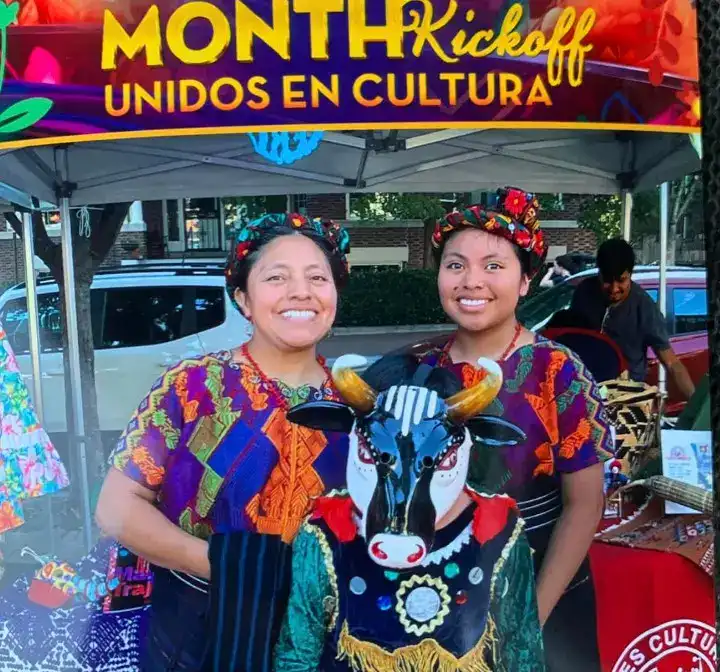 Vendedoras con vestimenta tradicional en una feria cultural, promoviendo la diversidad y el talento en eventos culturales latinoamericanos. Fondo con carpas y personas disfrutando del festival. – SoyMigrante.com REVISTA