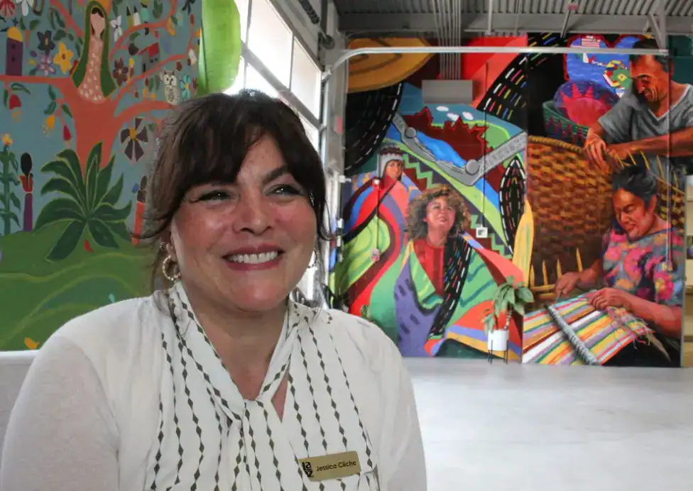 Colaboradora de SoyMigrante en evento cultural, con mural colorido de fondo que representa la riqueza cultural y tradición de comunidades migrantes en América Latina. – SoyMigrante.com REVISTA