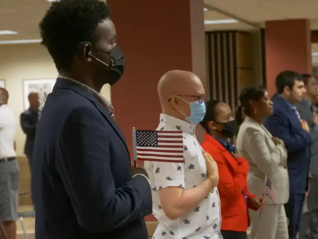 Manos sobre el corazón en acto cívico con banderas de EE.UU. y participación de diversas comunidades migrantes en evento oficial. – SoyMigrante.com REVISTA