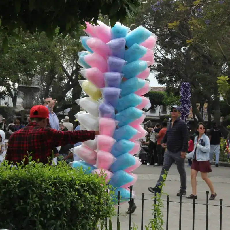 Bastón de algodón de azúcar multicolor en el parque, rodeado de personas disfrutando del evento y la feria, símbolo tradicional de dulzura y celebraciones populares en México. – SoyMigrante.com REVISTA