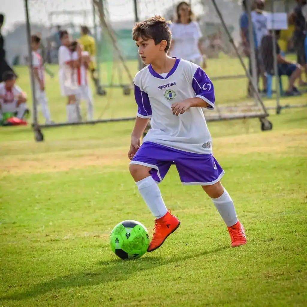 Niño jugando al fútbol en un campo al aire libre durante un partido infantil de deporte. – SoyMigrante.com REVISTA
