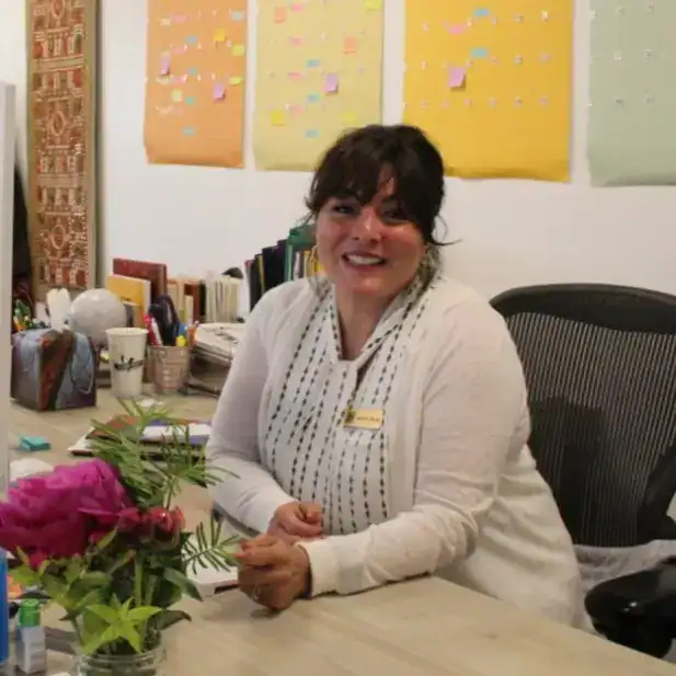 Vendedora sonriendo en oficina con fondo de notas adhesivas de colores, rodeada de libros y material de oficina, mostrando un ambiente laboral acogedor y amigable. – SoyMigrante.com REVISTA