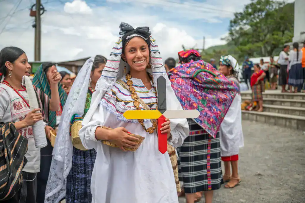 Primer plano de una mujer indígena vestida con traje tradicional, sonriendo y sosteniendo un símbolo de paz, en una celebración cultural en un pueblo de México. – SoyMigrante.com REVISTA