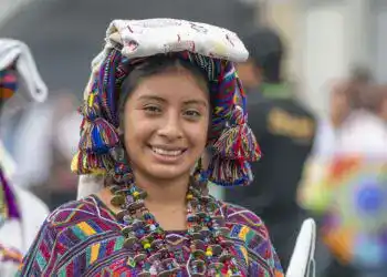 Vendedora indígena con traje tradicional colorido y accesorios en feria cultural en México, promoviendo identidad y cultura migrante.