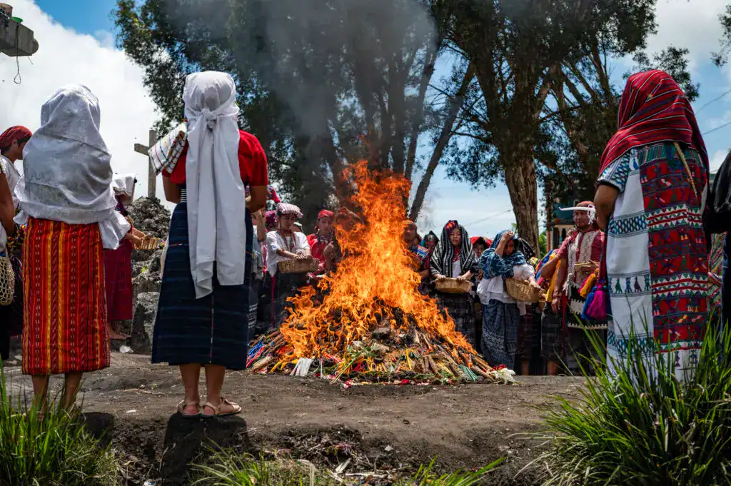 Centena de personas participando en ceremonia indígena con gran fuego, vestimenta tradicional y actos culturales en un entorno natural con árboles y cielo azul. – SoyMigrante.com REVISTA