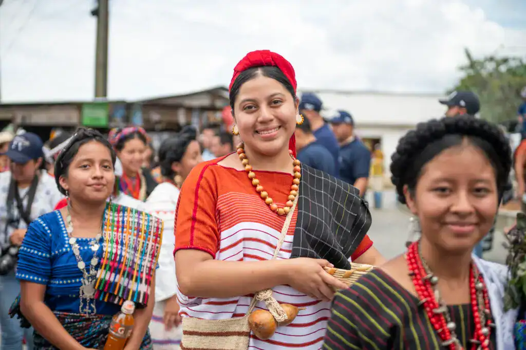 Coloridas mujeres indígenas en festividad tradicional con trajes típicos y accesorios culturales en una celebración en exteriores. – SoyMigrante.com REVISTA