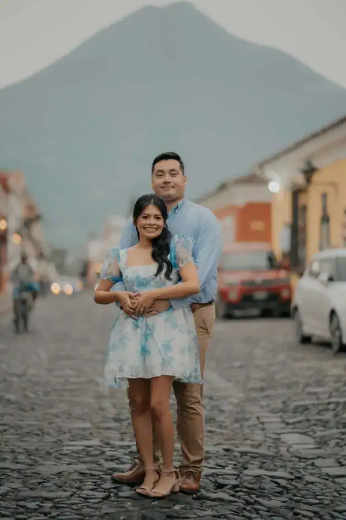 Vibrante pareja disfrutando de una caminata en una calle empedrada con el volcán de fondo, resaltando la belleza de Guatemala y su cultura en la revista SoyMigrante.com.