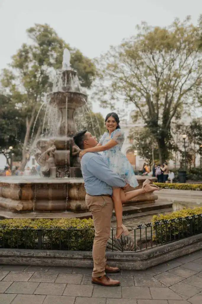 Chico sostiene a su novia en un parque con fuente, mostrando amor y felicidad, en un entorno urbano y natural, ideal para artículos sobre migrantes, amor y cultura en la revista SoyMigrante.com.