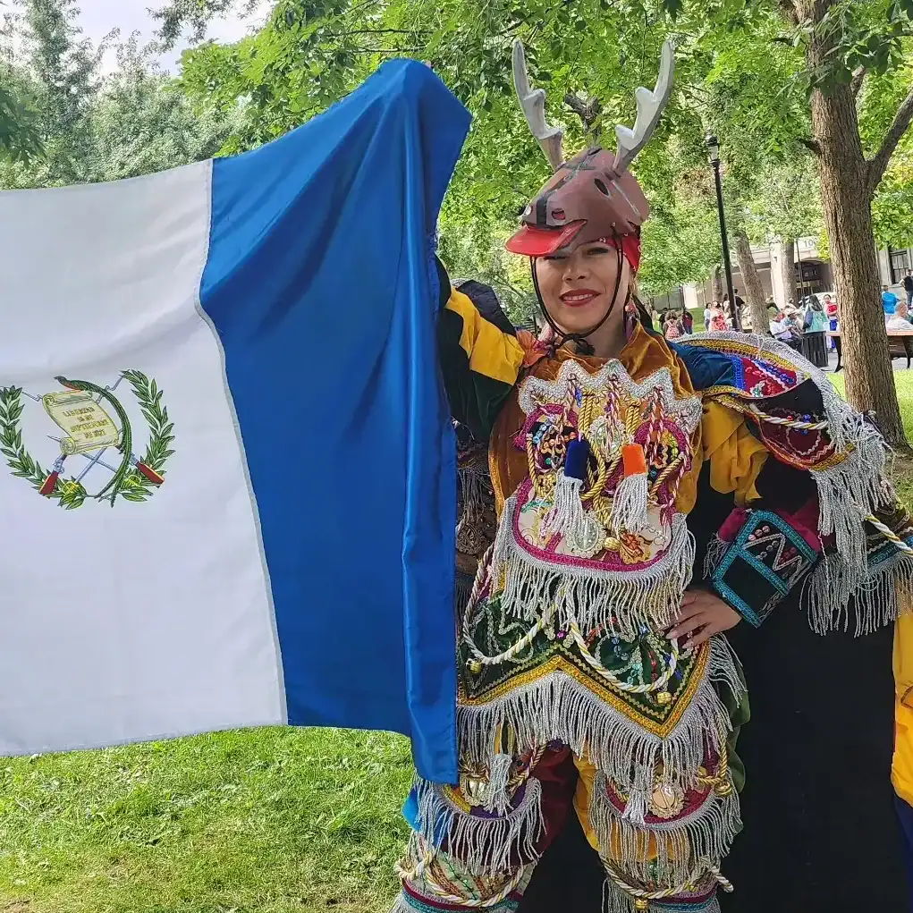 Rostro de mujer vestida con traje tradicional y casco de ciervo, mostrando la cultura indígena en un evento al aire libre con árboles y gente en el fondo.