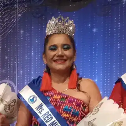 Joven con corona y banda de reina, en concurso de belleza cultural, luciendo vestimenta colorida y sonrisa radiante. – SoyMigrante.com REVISTA