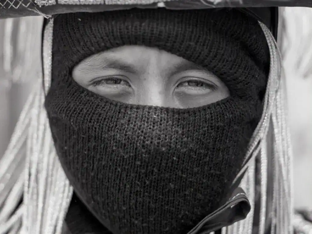 Rostro de un joven migrante con máscara negra y capucha en primer plano, en un contexto que refleja historias de migración y resiliencia.