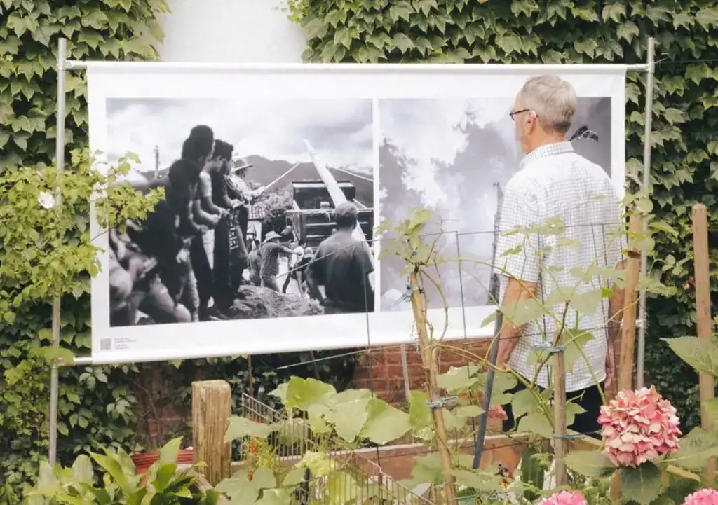 Fotografía de una exposición fotográfica sobre migrantes en un entorno al aire libre, con un hombre observando una imagen en la pared, rodeado de plantas y flores, promoviendo sensibilización y conciencia sobre migración.