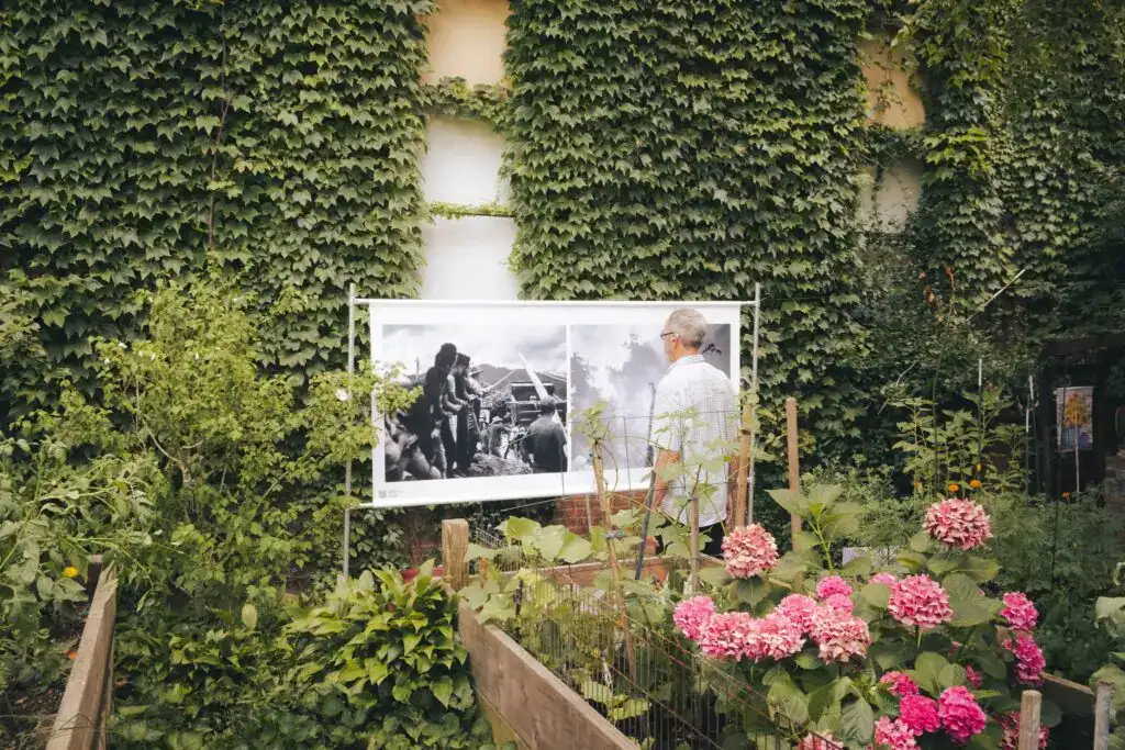 Fotografía de una exposición fotográfica en un jardín con muchas plantas y flores, resaltando la participación comunitaria en temas sociales y migrantes, en el contexto de SoyMigrante.com REVISTA.
