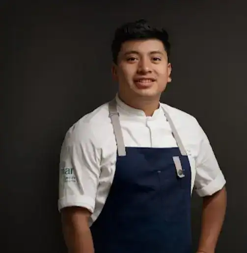 Joven chef mexicano sonriendo con delantal en fondo negro, promoviendo gastronomía y cultura en SoyMigrante.com Revista para migrantes y comunidades hispanas. – SoyMigrante.com REVISTA