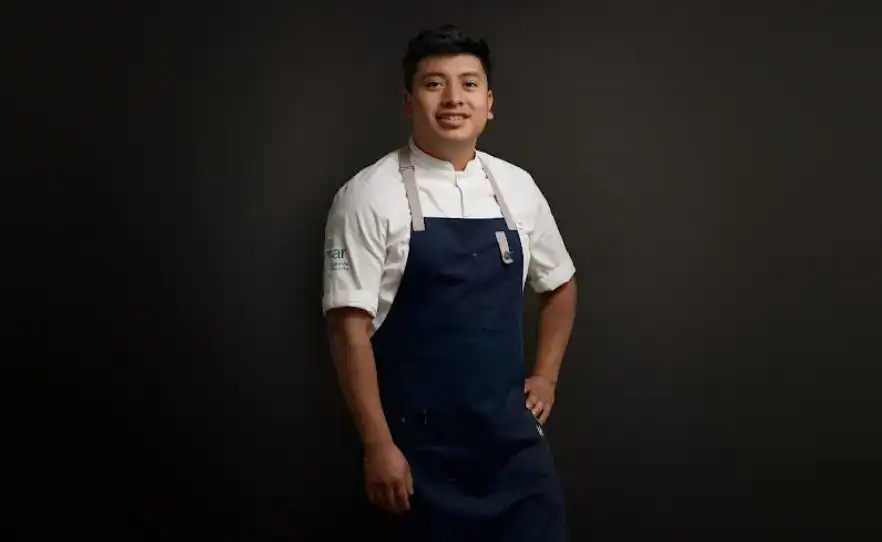 Mano de chef joven con uniforme en fondo negro, representación de inmigrantes en la gastronomía, temática de diversidad, integración y cultura en la comunidad migrante. – SoyMigrante.com REVISTA