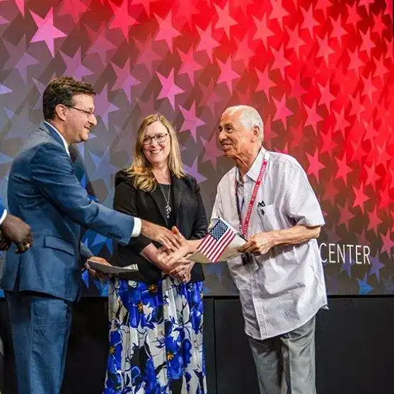 Recibiendo reconocimiento en un evento, tres personas en un escenario con fondo de estrellas rojas y moradas, un hombre en traje azul, una mujer con vestido floral y un hombre mayor con camiseta blanca y bandera de EE.UU. – SoyMigrante.com REVISTA
