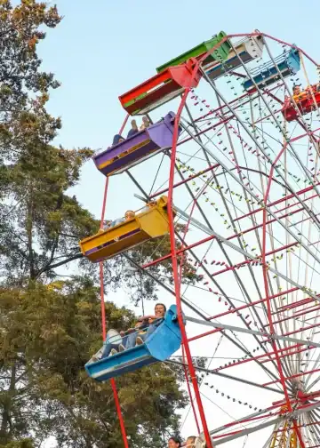 Parque de diversiones con rueda de la fortuna colorida y personas disfrutando en los asientos, rodeado de árboles verdes, en un día soleado. – SoyMigrante.com REVISTA