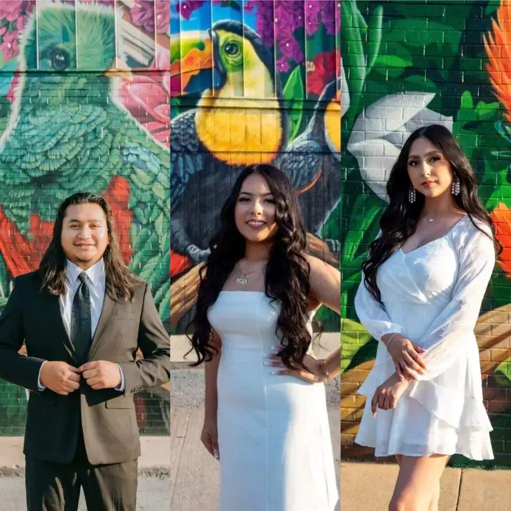Retrato de tres jóvenes en ropa formal frente a un mural colorido con aves y plantas, celebrando la cultura y diversidad en un evento de migrantes, reflejando orgullo y identidad. – SoyMigrante.com REVISTA