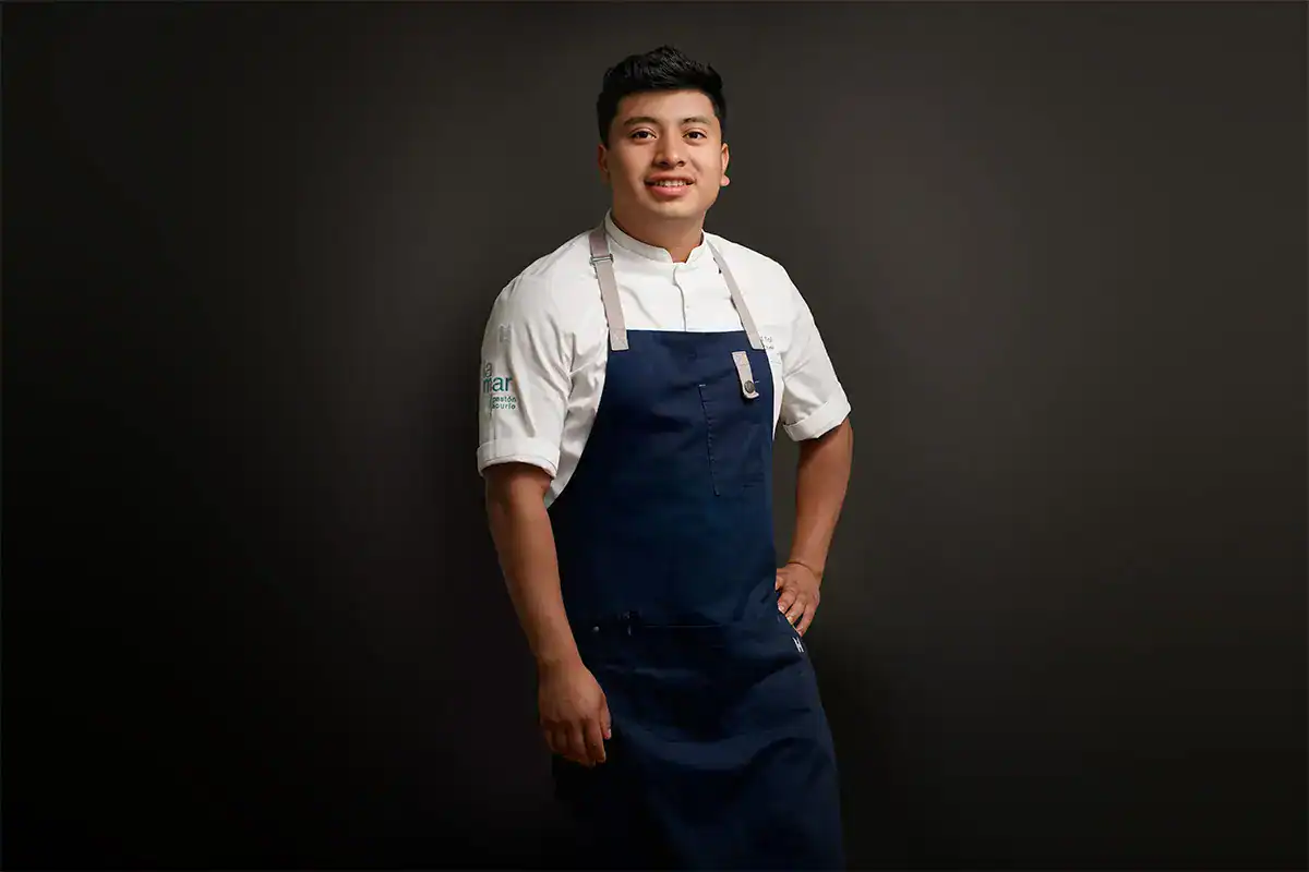 Chef Manuel Tol de Guatemala en Miami – SoyMigrante.com REVISTA Joven cocinero con uniforme profesional en un fondo oscuro, promoviendo la cultura migrante y el talento en la gastronomía.