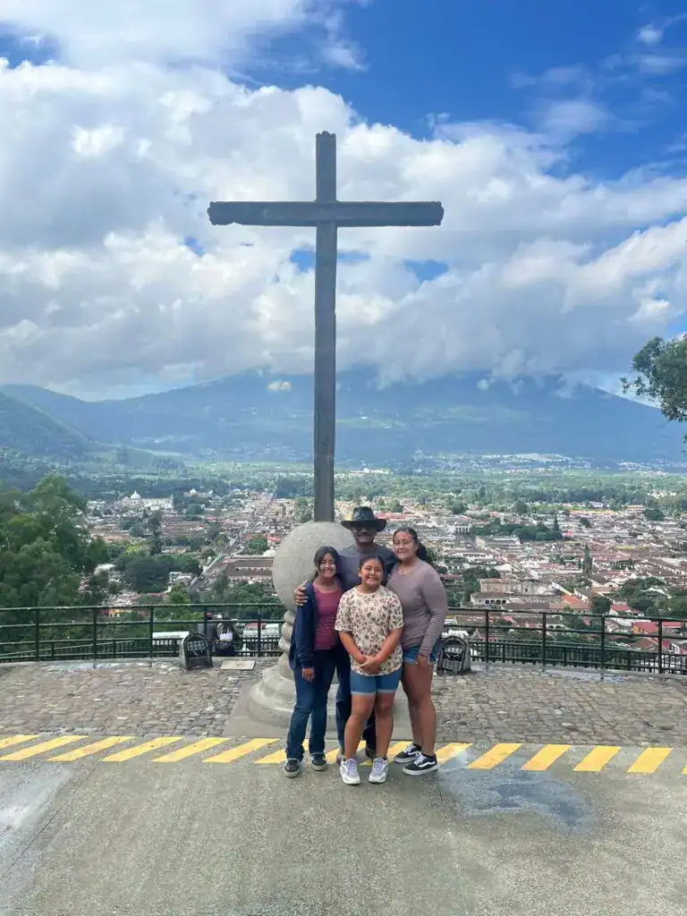 Cruz de madera en una colina con vista panorámica de la ciudad y montañas, en un día despejado, fotografía familiar en Guatemala. – SoyMigrante.com REVISTA