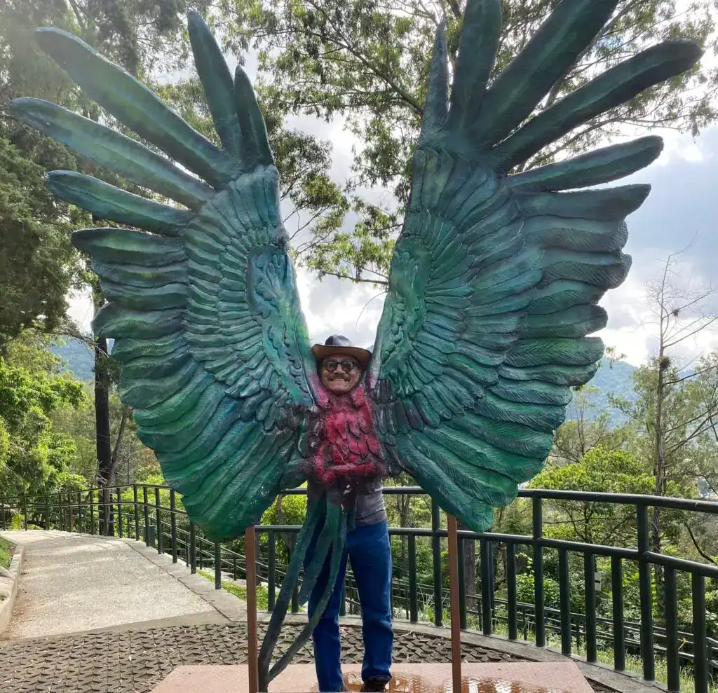 Vendedor en celebración con gran disfraz de alas de ave en un entorno natural, mostrando cultura y expresiones artísticas en espacios públicos. – SoyMigrante.com REVISTA